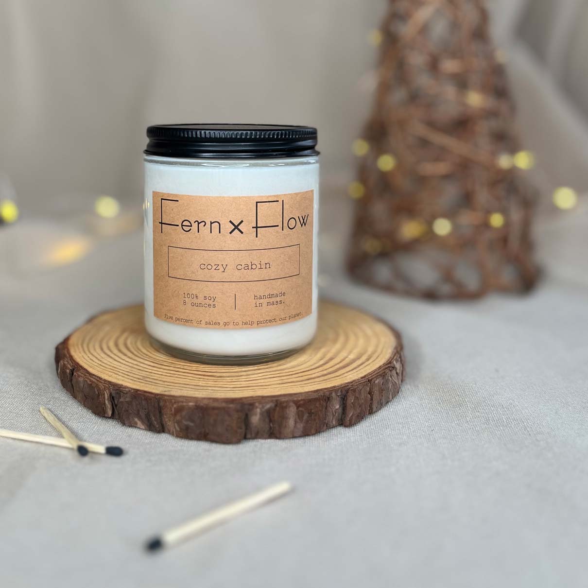 Fern x Flow Cozy Cabin scented soy candle on a rustic wooden riser with black tipped safety matches in the foreground and white lights and a wicker Christmas tree in the background.