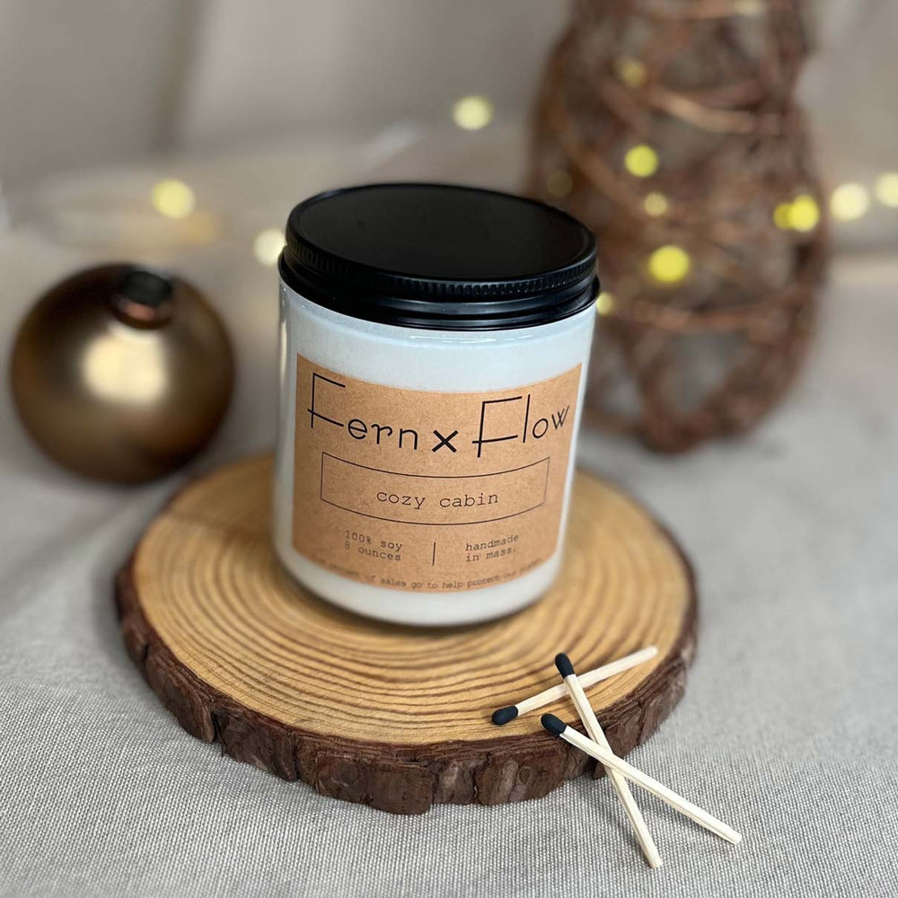 Fern x Flow Cozy Cabin holiday scented soy candle on a rustic wooden riser with black-tipped matches in the foreground and a wicker Christmas tree and golden ornament in the background.