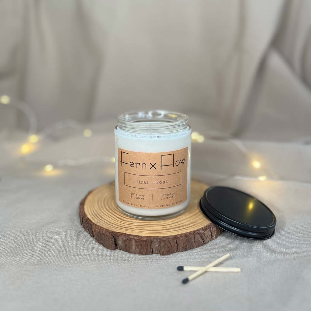 Fern x Flow eight-ounce First Frost scented soy candle on a rustic wooden riser with Christmas lights out-of-focus in the background.