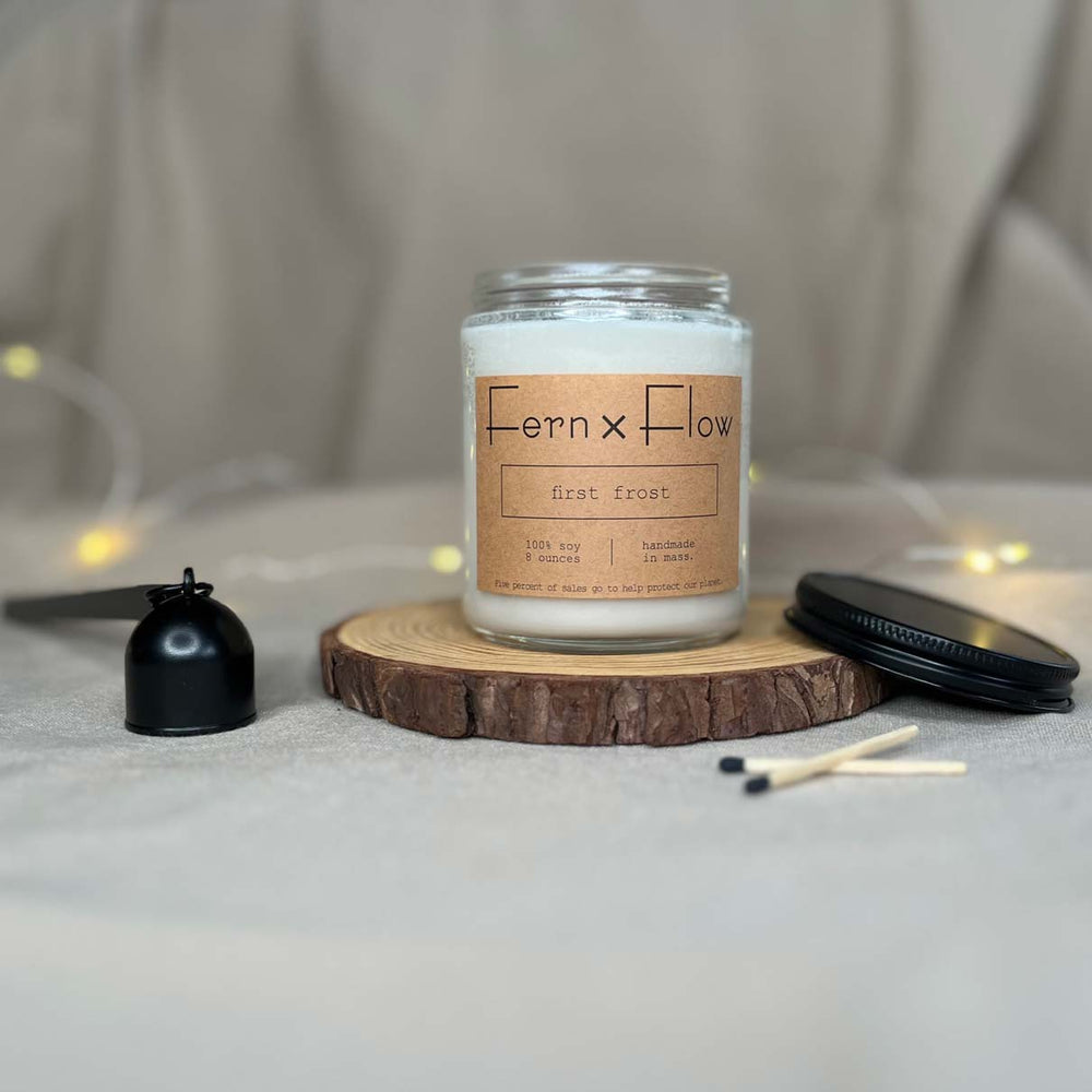 Fern x Flow eight-ounce First Frost scented soy candle on a rustic wooden riser with a black matte lid and candle snuffer on the sides.