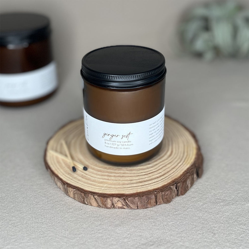 Fern x Flow 8oz Ginger Zest scented summer soy candle on a rustic wooden riser with a large air plant out-of-focus in the background.