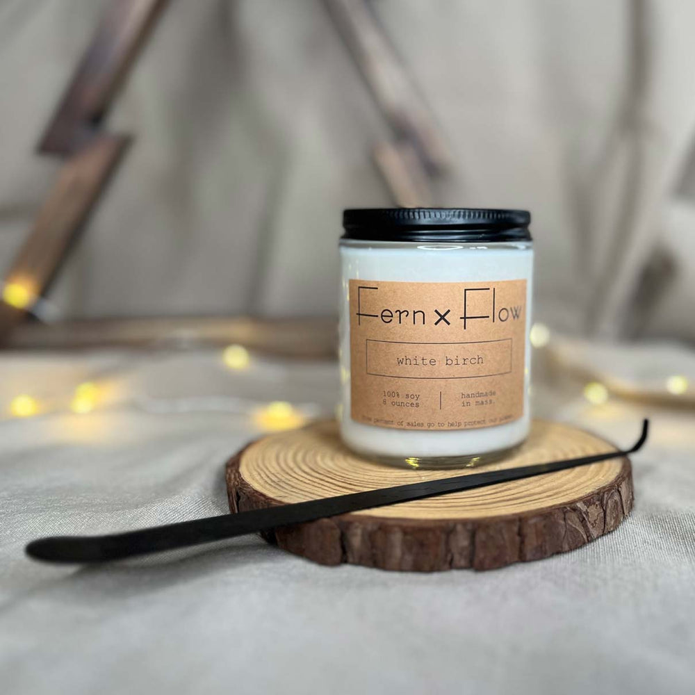 Fern x Flow eight-ounce White Birch scented holiday soy candle on a rustic wooden riser with a matte black wick dipper in the foreground and a wooden Christmas tree and white lights in the background.